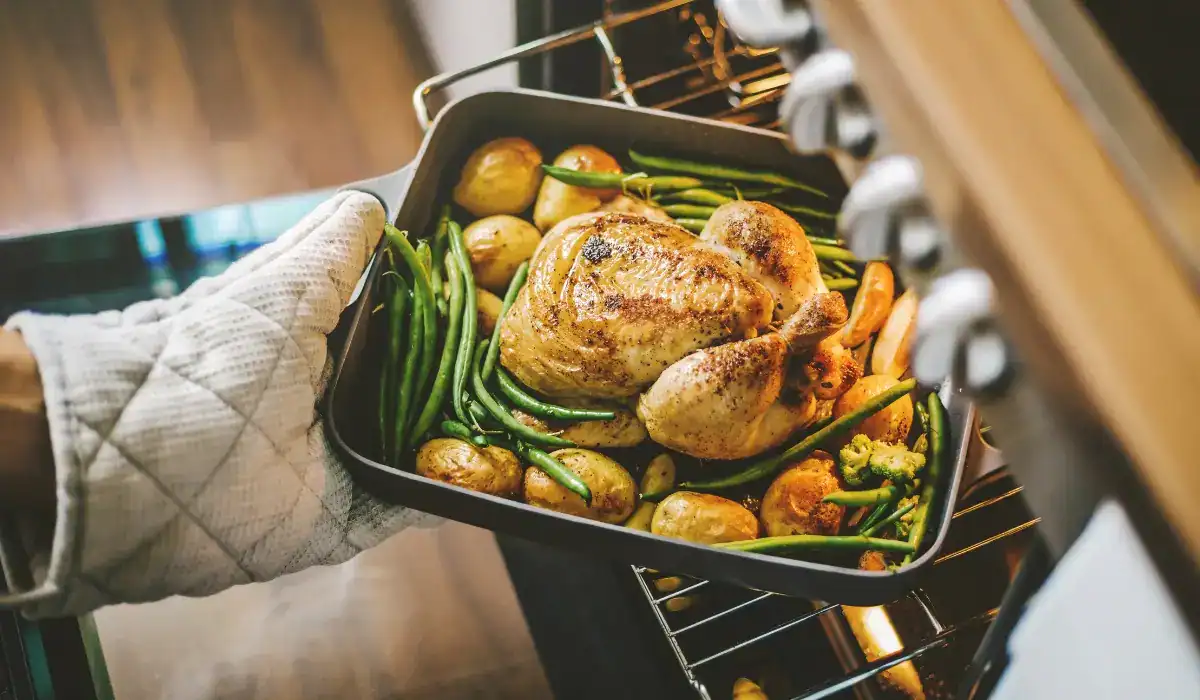 Roasted chicken and vegetables being pulled from a working oven during meal prep, showing appliances are ready for hosting.